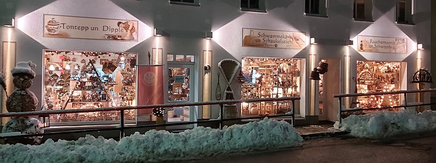 Die Aussenansicht unseres Geschäftes mit drei Schaufenstern im Winter ist effektvoll beleuchtet. Auf dem Gehweg stehen ein geflochener Schneemann etwa 1,5m groß und andere Dekorationen aus Weidenzweigen. 