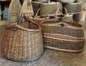 Zahlreiche Einkaufskörbe mit Wurzelholzgriff in dunkler ungeschälter Weide sind zu sehen. Die Körbe sind rund oder oval und haben teils Ornamente aus heller oder hellbrauner Weide eingeflochten.