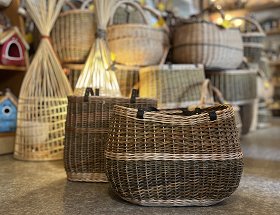 Zwei Hand- und Transportaschen mit Lederriemen füllen dieses Bild aus. Die Handtasche ist länglich und schmal. Die Transporttasche ist oval. Beide bestehen aus dunkler Weide, haben helle Streifen eingeflochten und sind zum Tragen in der Hand gedacht.