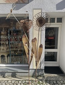 Links neben der Eingangstür zum Geschäft steht ein drittes Dekorationsobjekt auf dem Gehweg. Dieses zeigt etwa zwei Meter große, geflochtene Blumen mit unterschiedlichen Blütenkelchen und -blättern. Helle und dunkle Weidenzweige wurden dafür verwendet, wodurch sie einen schönen Kontrast liefern.