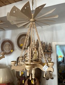 Eine hölzerne erzgebirgische Deckenpyramide hängt im Geschäft an einer dünnen Kette. Sechs filigran geschnittene Holzstäbe schaffen die Verbindung zwischen den Flügeln und dem unteren Teil der Pyramide. Geschnitzte Bäume und Figuren aus dem bäuerlichen Leben sind auf dem Drehteller zu sehen.