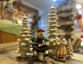 Drei geschnitzte Bäume und zwei traditionelle erzgebirgische Figuren sind hier als Einzelmotive dargestellt. Eine Figur zeigt einen alten Spanbaumschnitzer mit Pfeife auf einem Birkenstumpf sitzend. Die zweite Figur stellt eine Schwammefraa mit Korb und weitem, gelblichen Gewand dar.