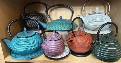 Sieben japanische Gussteekannen unter anderem in türkis, lila weiß und rot stehen in einem Schrank. Diese uralte Handwerkskunst ist sehr filigran gearbeitet, hat jeweils einen ausladenden Ausgießer und einen rundlichen Henkel. Die Kannen stehen außerdem auf Untersetzern.