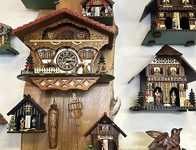 Zahlreiche Motive von Schwarzwälder Kuckucksuhren und Wetterhäuser sind auf diesem Bild dargestellt. Details sind auf der linken Seite gut zu erkennen. Hier wird ein typisch Schwarzwälder Haus mit rotem Dach und hölzernem Balkon dargestellt. Unter dem Balkon ist ein großes Wasserrad und ein Schmied mit Hammer und Amboss zu sehen. Dazwischen sieht man das große Ziffernblatt der Uhr, unter dem die schweren Gewichte und das Pendel hängen.