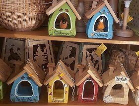 Sechs bunte, handbemalte Lärchenvogelhäuser stehen auf zwei Regalböden. Leuchtende Farben und kesse Bezeichnungen, wie Zwitscherstube oder Liebesnest zeichnen diese außergewöhnlichen, krummen und schiefen Häuschen aus.